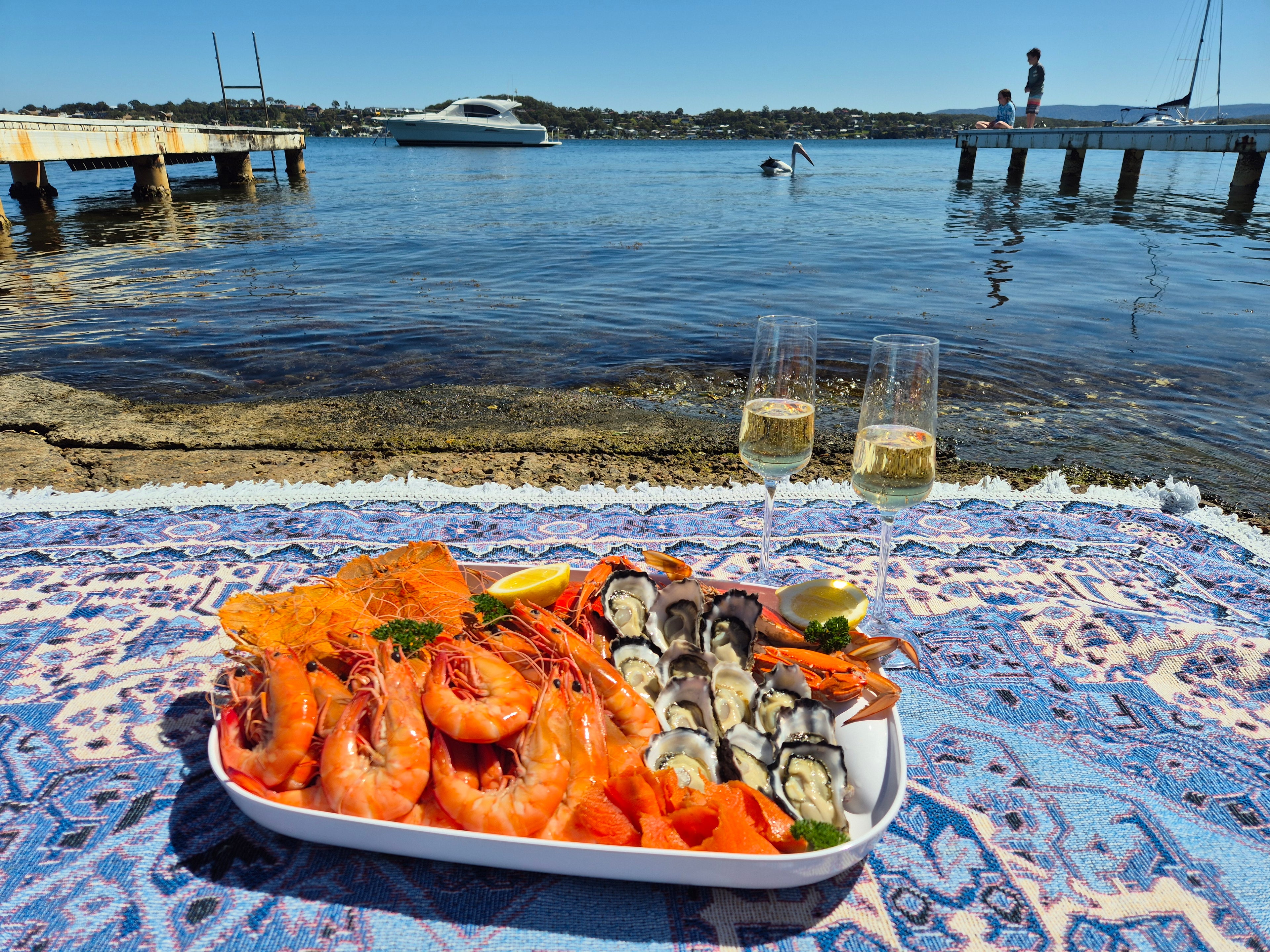 Platter of seafood and oysters with two glasses of champagne on a patterned tablecloth by a waterfront.